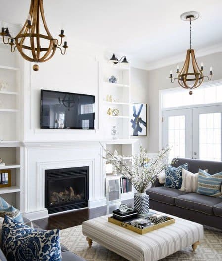 Bright living room with white built-in bookcases, a cozy fireplace, wooden chandeliers, and a tufted ottoman centerpiece