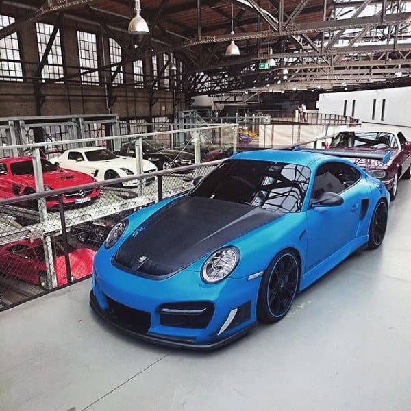 A spacious industrial-style garage showcasing a vibrant blue luxury car on an elevated mezzanine with classic and modern vehicles below.