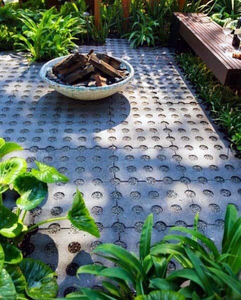 A small patio features a concrete surface, a fire pit with stacked wood, and surrounding lush green plants