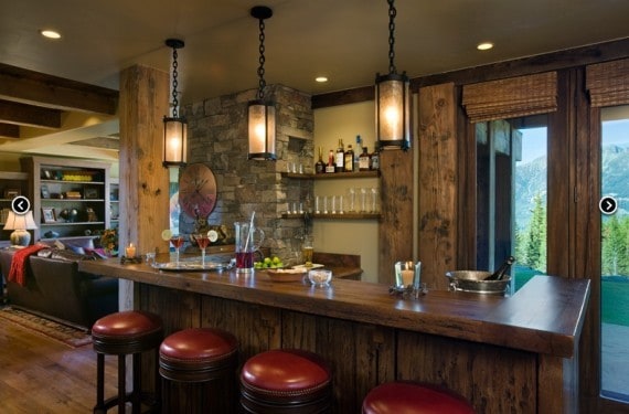 Cozy rustic bar with wooden accents, stone wall, and red cushioned stools, overlooking lush greenery outside