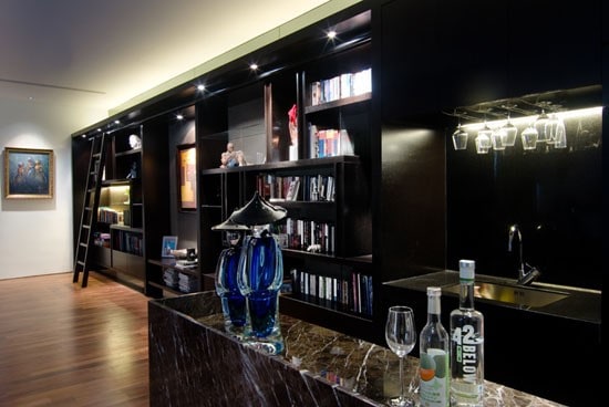 Modern bar with dark shelves, a ladder, and a marble counter featuring bottles, glasses, and blue decor