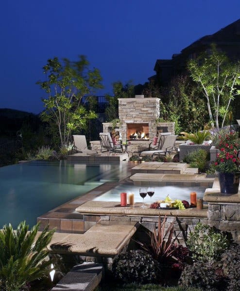 Backyard with infinity pool, stone fireplace, hot tub, and cozy nighttime ambiance.