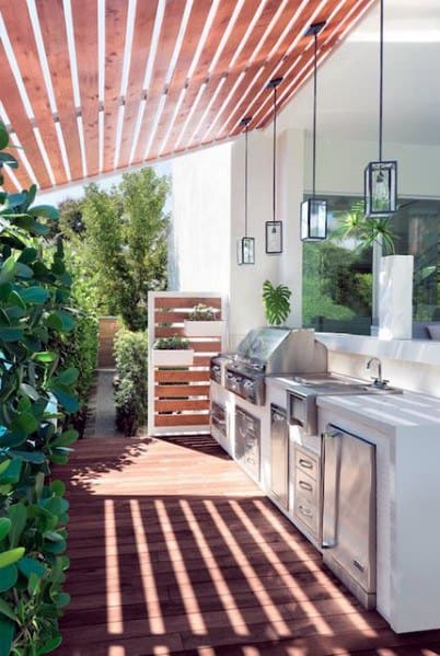 Outdoor kitchen with built-in grill, modern appliances, wood deck, pergola, greenery; hanging lanterns add charm