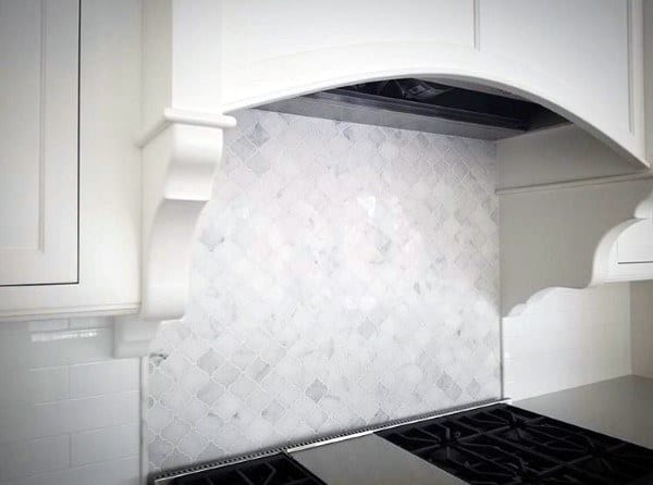 White mosaic tile backsplash with custom range hood and stainless steel gas stovetop.
