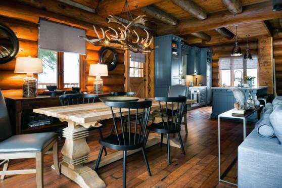 Modern log cabin dining area with antler chandelier, rustic wooden table, and cozy kitchen.