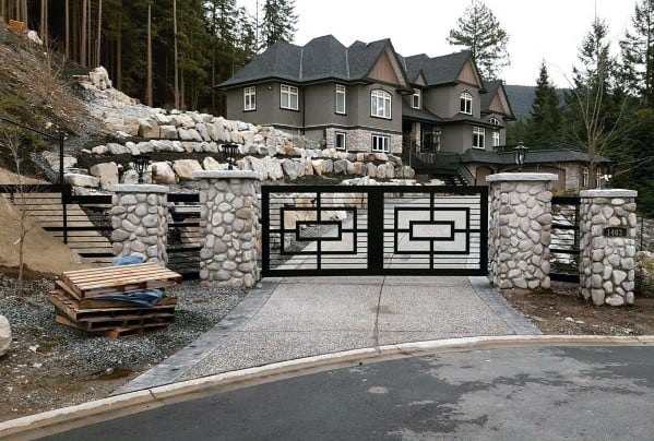Black metal driveway gate with geometric patterns between stone pillars.