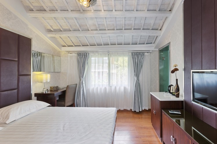 A luxury modern hotel room with an unfinished white ceiling, a cozy bed, a wooden floor, and large windows allowing natural light, creating a clean, open, and tranquil ambiance