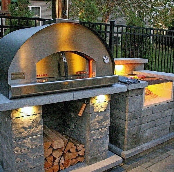 Outdoor pizza oven setup with firewood storage, warmly lit on a stone patio with lush greenery surrounding