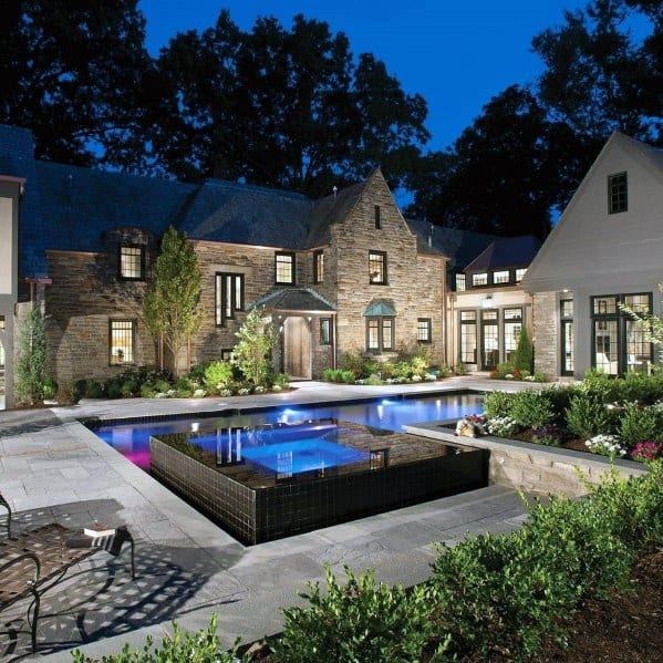 Stone mansion with lit windows, a modern pool, and lush landscaping at dusk