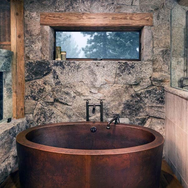 Rustic bathroom with stone walls and a round copper tub beneath a small horizontal window