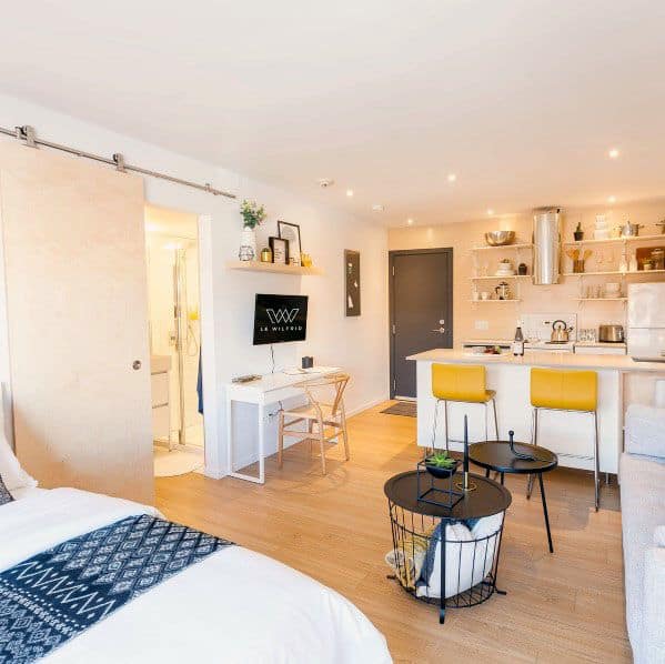 Bright studio with open kitchen, cozy bed, workspace, and vibrant yellow bar stools.