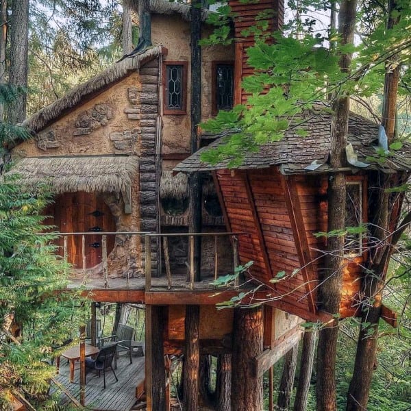 A rustic treehouse surrounded by lush green forest with a small wooden deck and chairs
