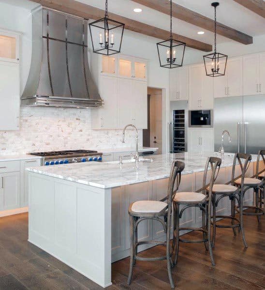 White kitchen with marble island, exposed beams, pendant lighting, and metal range hood.