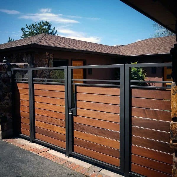 Modern wooden and metal gate with horizontal panels, leading to a house entrance under a clear blue sky