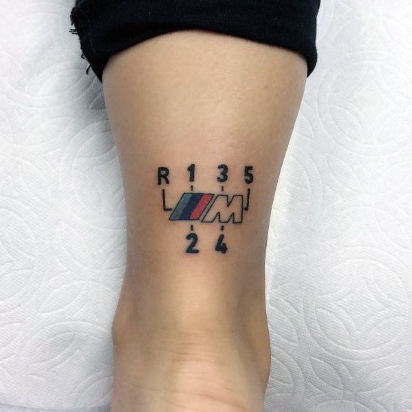 Minimalist BMW M gear shift tattoo on ankle, featuring the M logo and classic shift pattern for a sleek automotive-inspired design.