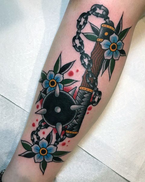 Vibrant mace tattoo with a spiked ball, chain, and wooden handle, accented by blue flowers and red splashes, symbolizing strength and beauty.