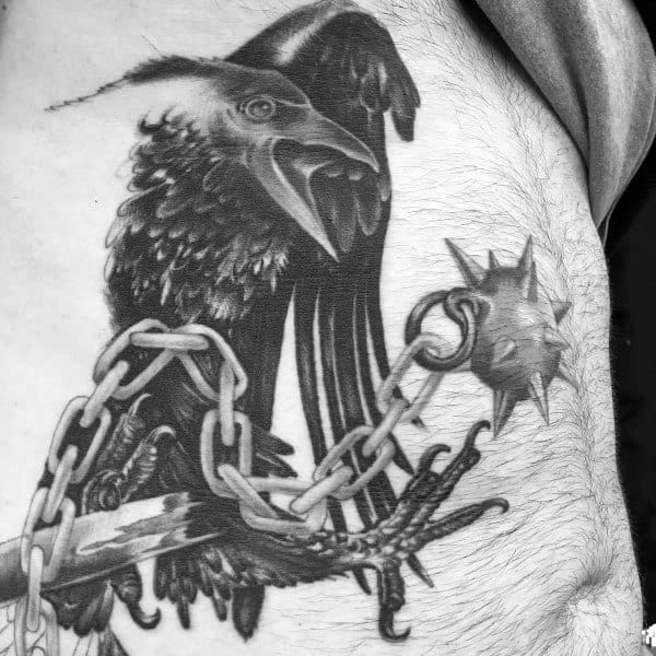 Detailed black and gray tattoo of a raven gripping a spiked mace with chains, symbolizing power, mystery, and battle in a striking design.