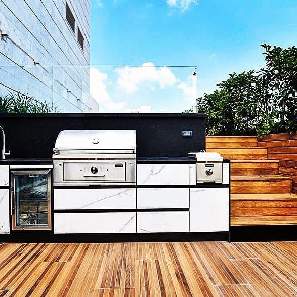 Modern outdoor kitchen with a built-in grill, marble countertop, and wooden deck steps, surrounded by greenery