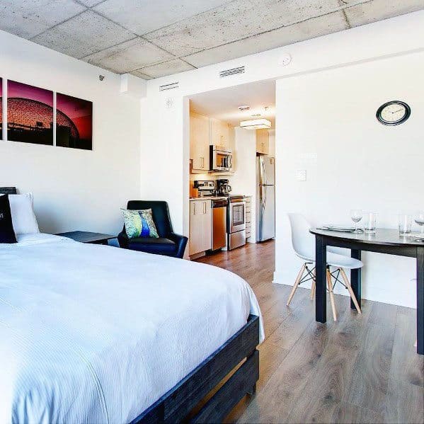 Modern studio with exposed ceiling, cozy bed, small dining area, and sleek kitchen.