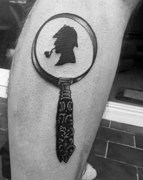 Tattoo of a magnifying glass with Sherlock Holmes’ silhouette inside, featuring bold black ink and an ornate handle for a detective-inspired design.