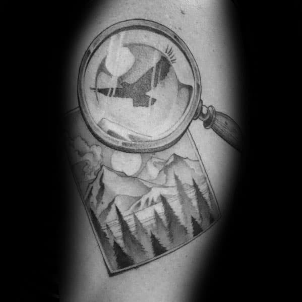 Tattoo of a magnifying glass over a mountain landscape photo, revealing a soaring bird, symbolizing adventure and discovery in a detailed black-and-gray design.