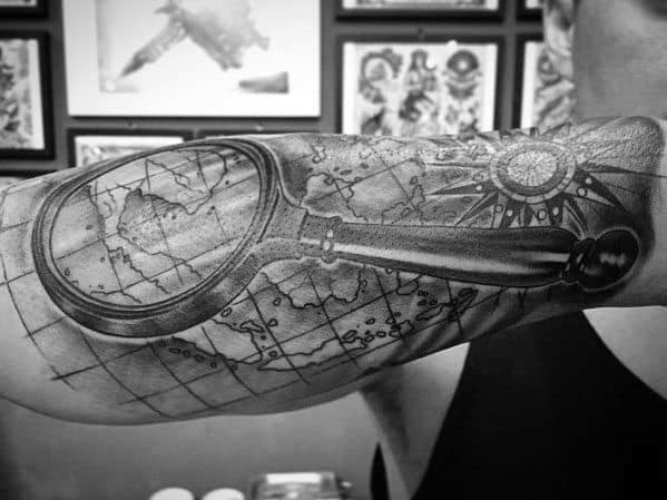 Tattoo of a magnifying glass over a detailed world map with a compass rose, creating a bold and realistic travel-inspired forearm design.
