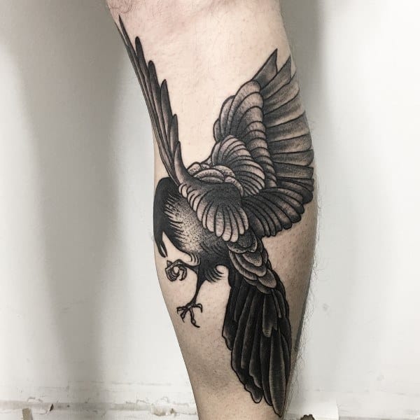 Black and gray magpie tattoo on the leg, featuring the bird in mid-flight with intricate feather details and shading, holding a small object in its claws.