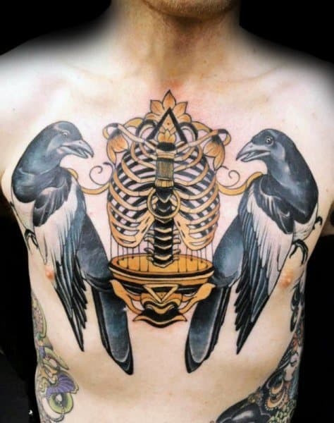 Intricate chest tattoo featuring two magpies perched on either side of a golden ribcage and cage-like design, blending realism with ornamental elements.