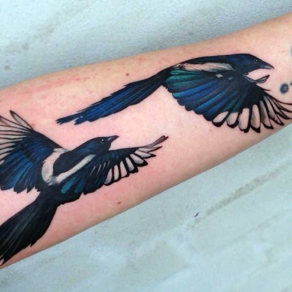 Two magpies in flight tattoo on forearm, featuring bold black, white, and blue feathers with dynamic wing positions for a lively design.