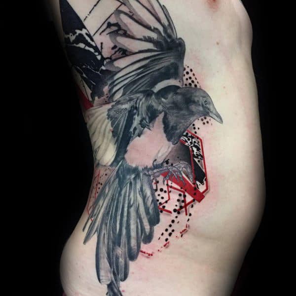 Black and gray magpie tattoo on the ribs, featuring a dynamic bird in flight with abstract red and geometric elements for a bold, modern look.
