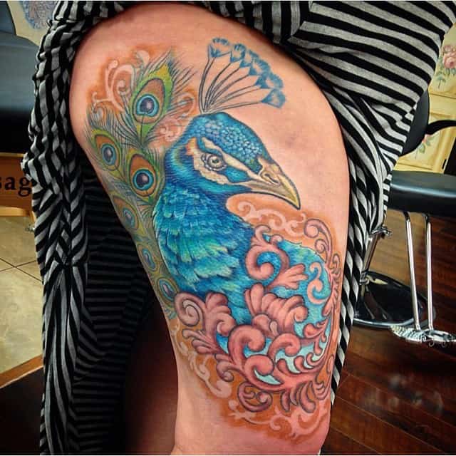 Colorful peacock tattoo with blue and green feathers, accented by soft pink and orange swirls, placed on the upper thigh.