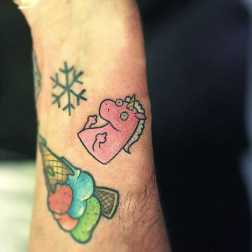 A colorful tattoo of a pink cartoon unicorn with a goofy expression, placed near an ice cream cone and snowflake design on the wrist.