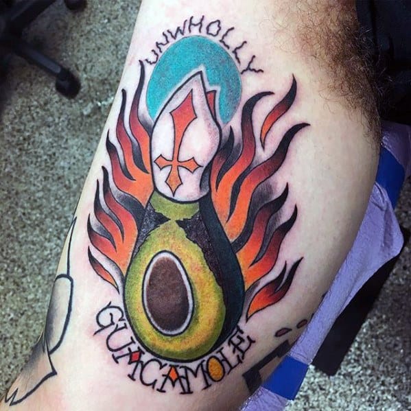 Bold avocado tattoo with a bishop hat, flames, and the words “Unwholly Guacamole,” mixing humor, religion, and vibrant color.