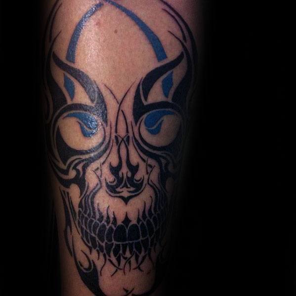 Tribal skull tattoo with bold black and blue ink, featuring sharp symmetrical patterns and piercing eyes on the forearm.