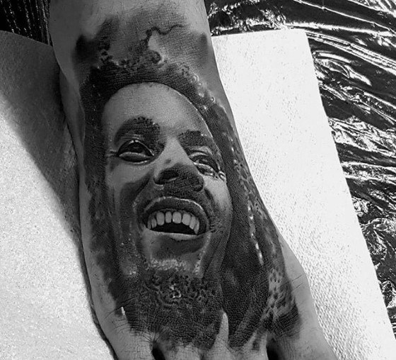 Realistic black and gray foot tattoo of Bob Marley smiling, with detailed facial shading and textured dreadlocks.