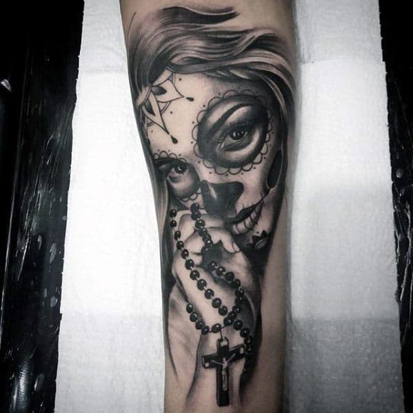 La Catrina tattoo of a woman with sugar skull makeup holding a rosary, featuring intense eyes and fine black and grey shading.