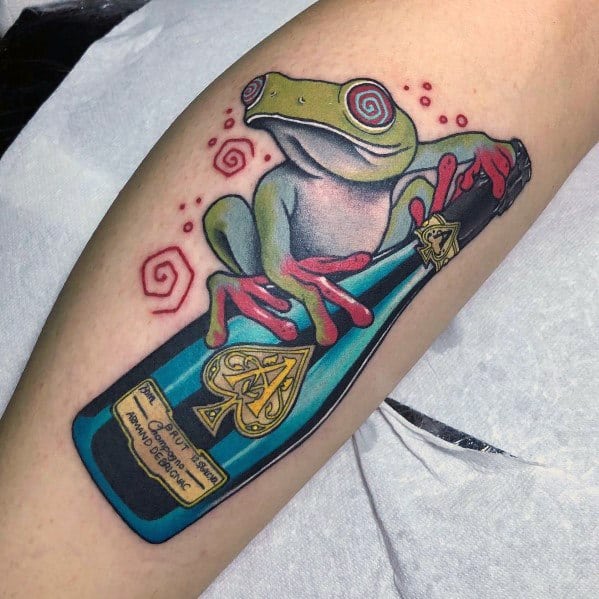 Tattoo of a green frog with red eyes and limbs sitting on a bottle of champagne, surrounded by swirling patterns.