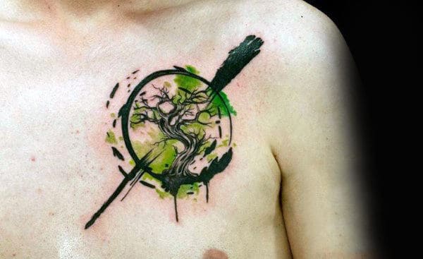 Abstract watercolor chest tattoo of a twisted tree inside a bold, black circular brushstroke, accented with green splashes for a dynamic artistic effect.