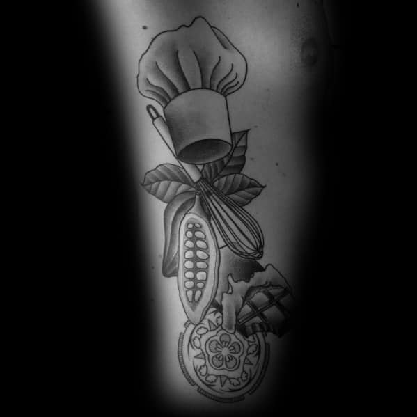 This tattoo features a detailed black-and-white culinary-themed design, including a chef’s hat, cocoa pod, whisk, chocolate, and a decorative cookie.