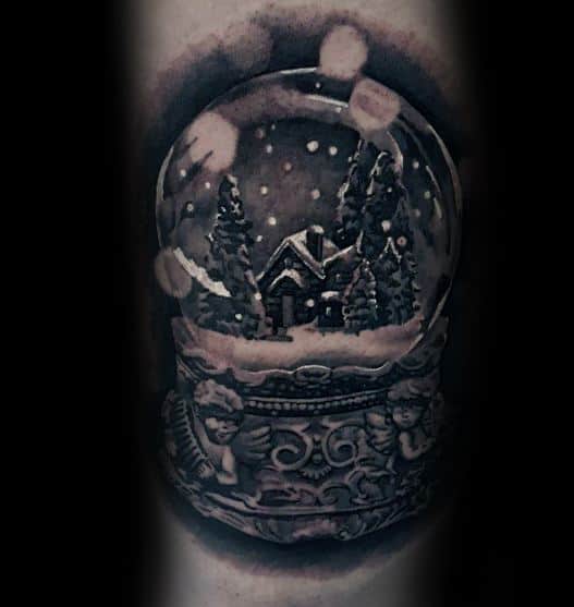 Black and grey Christmas tattoo of a detailed snow globe featuring a cozy cabin, snow-covered trees, falling snowflakes, and ornate angel carvings on the base.