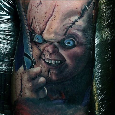 Realistic Chucky tattoo with vivid details, featuring his sinister grin, stitched face, glowing blue eyes, and a sharp knife for a chilling horror look.