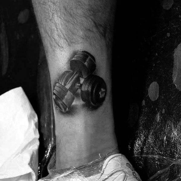 Realistic ankle tattoo of crossed dumbbells in black and grey, representing strength, dedication, and a grounded fitness lifestyle.
