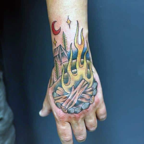 Colorful hand tattoo with a campfire, pine trees, mountains, crescent moon, and stars in a stylized outdoor scene.