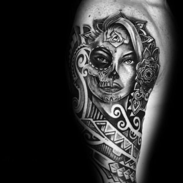 La Catrina tattoo featuring a half-skull woman with tribal patterns, a rose on her head, and bold black geometric detailing.