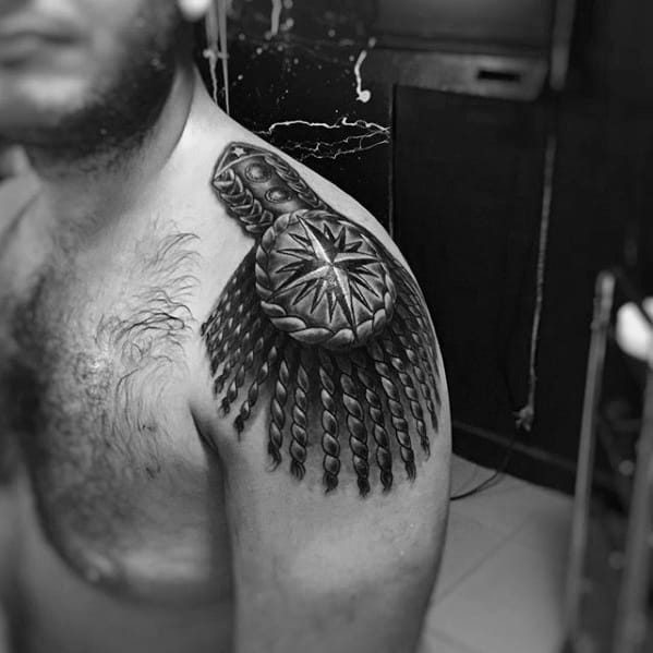 Bold epaulette tattoo with a large compass rose centerpiece, braided border, and thick twisted tassels cascading over the shoulder.