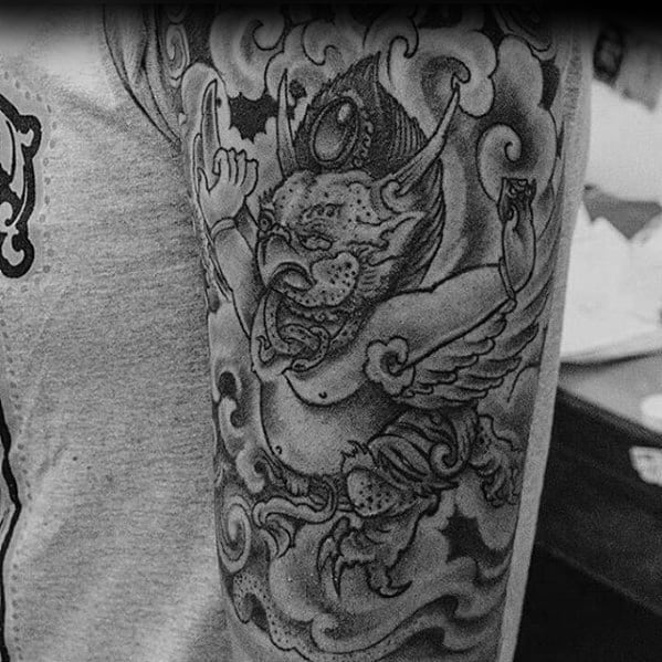 Garuda tattoo design showing a winged mythical creature in motion with fierce features and cloudy background on the upper arm.
