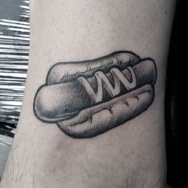 Tattoo of a black and grey hot dog featuring a sausage with mustard in a soft bun on the ankle.