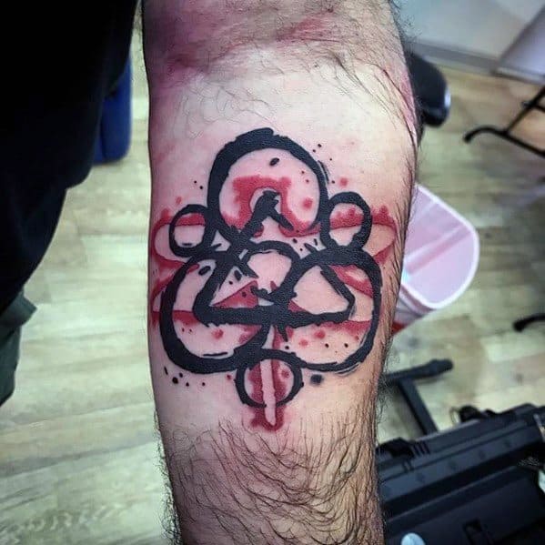 Coheed and Cambria Keywork tattoo on the inner arm with bold black ink and red watercolor splashes for a vivid, rebellious look.