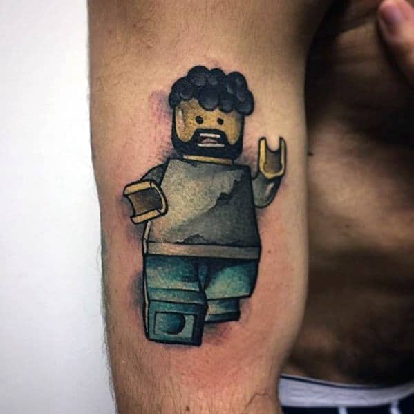 Tattoo of a custom Lego minifigure with curly hair, beard, casual clothes, and raised hand, done in a watercolor-style design on the bicep.