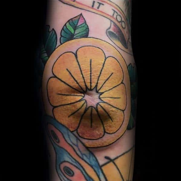 Bold and vibrant tattoo of a lemon slice with thick black outlines and orange-yellow shading, surrounded by green leaves and other elements.